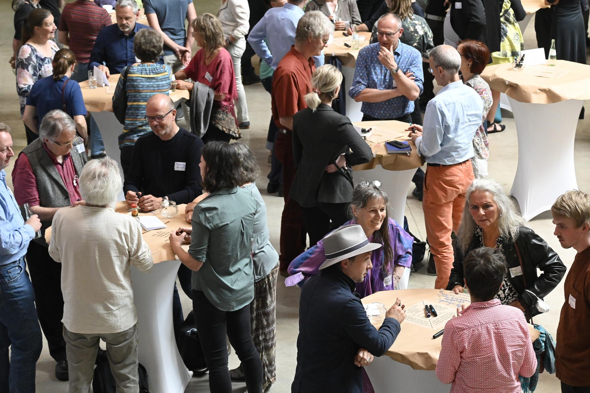 Mehrere Personengruppen stehen um Stehtische herum und sprechen miteinander.