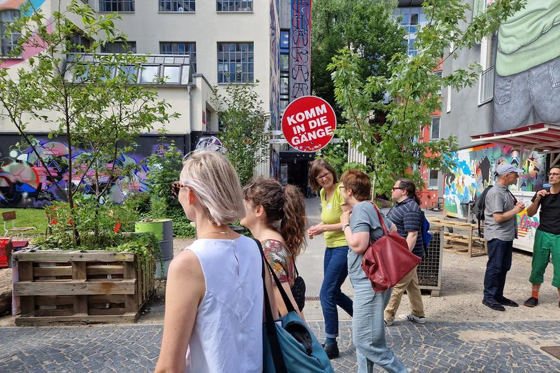 Urbane Straßenszene mit mehreren Personengruppen, die vorbeigehen und sich unterhalten. Im Hintergrund ein rotes rundes Schild mit der Aufschrift "Komm in die Gänge"
