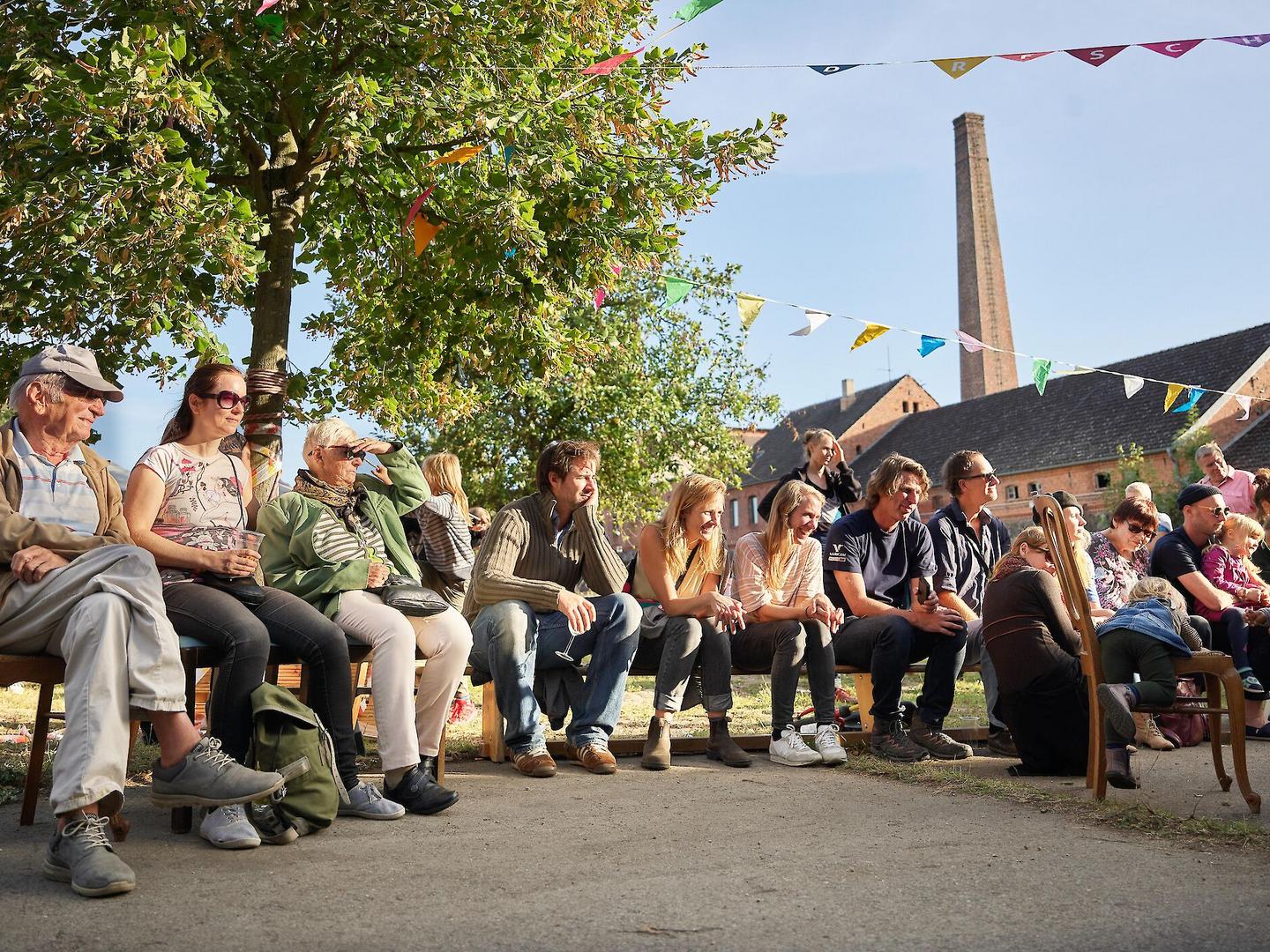 Viele Menschen unterschiedlichen Alters sitzen auf einer Bank im freien unter Wimpelketten in der Sonne.