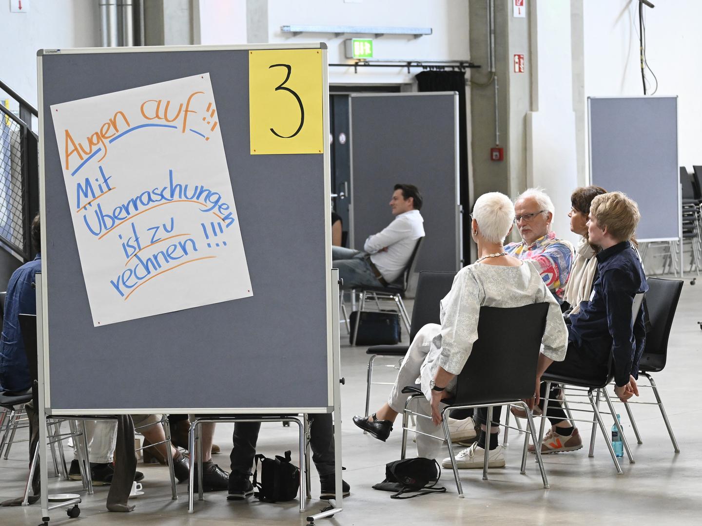 Eine Gruppe von Menschen sitzt im Kreis in einer Besprechung. Im Vordergrund eine Stellwand mit der Aufschrift: "Augen auf! Mit Überraschungen ist zu rechnen!"