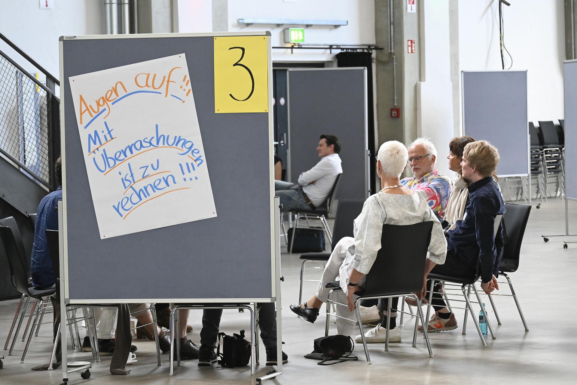 Eine Gruppe von Menschen sitzt im Kreis in einer Besprechung. Im Vordergrund eine Stellwand mit der Aufschrift: "Augen auf! Mit Überraschungen ist zu rechnen!"